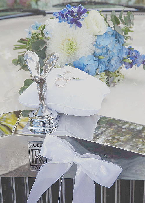 Front of a Rolls Royce with wedding ring on a cushion an white and blue flowers